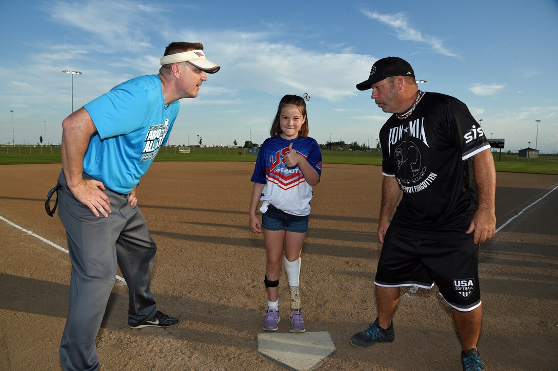 Tech. Sgt. Erik Clemenson, of the 119th Security Forces Squadron, left, and retired U.S. Air Force Chief Master Sgt. Randall Raper watch Annie Hanning flip a coin for home field designation between the Fargo area law enforcement all-stars and the USA Patriots (formerly known as the wounded warrior amputee softball team) at the softball complex north of Fargo, N.D., June 14, 2019.