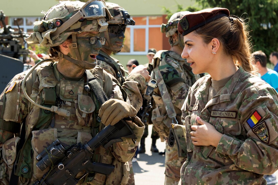 A U.S. soldier carrying a rifle and wearing a uniform and camouflage face paint talks to a Romanian soldier.