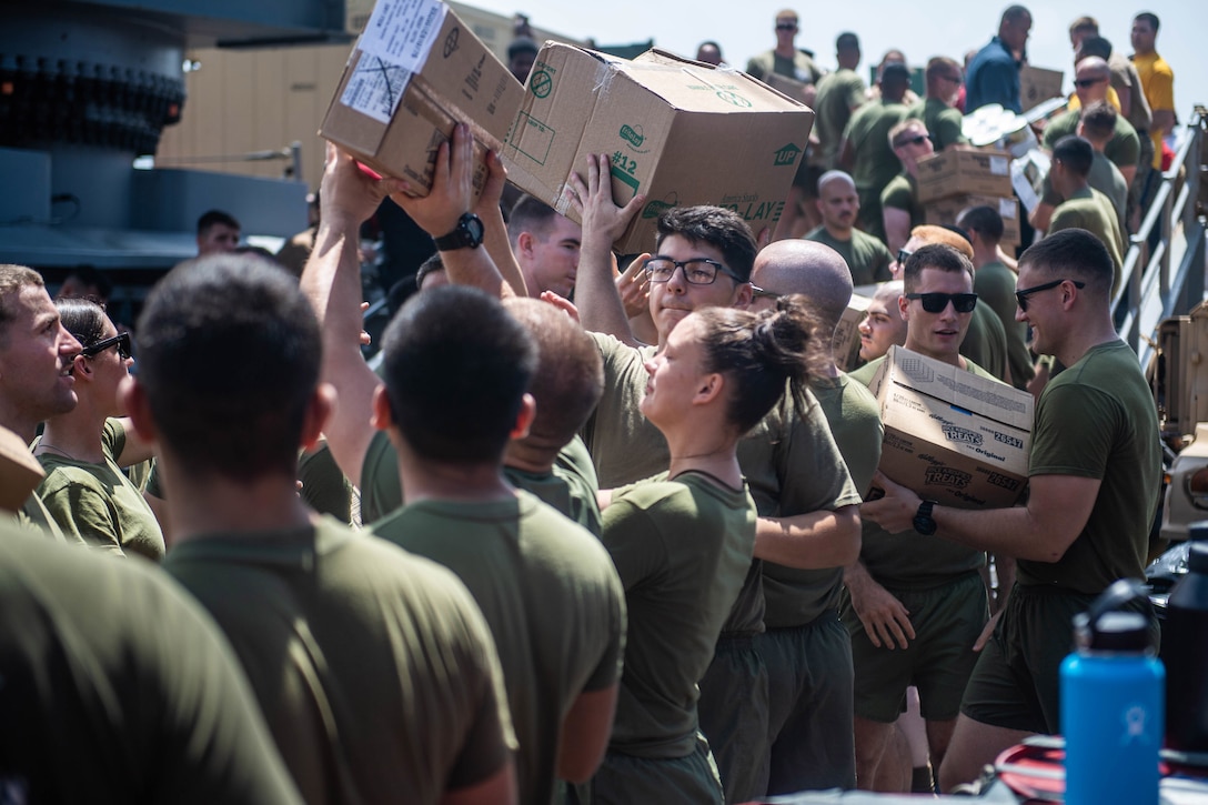Sailors and Marines line up and pass boxes of supplies to one another aboard a ship.
