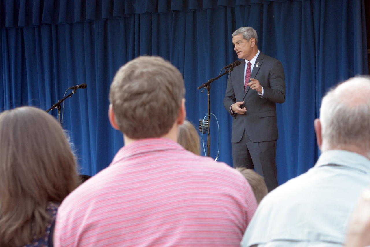 A man speaks from behind a microphone. In the background is a blue curtain. In the foreground are the backs of the heads of the audience.