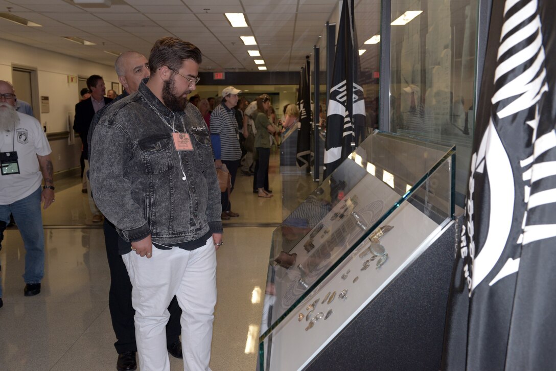 A man looks at a museum-like exhibit in a large hallway. In the foreground looms a black POW/MIA flag.