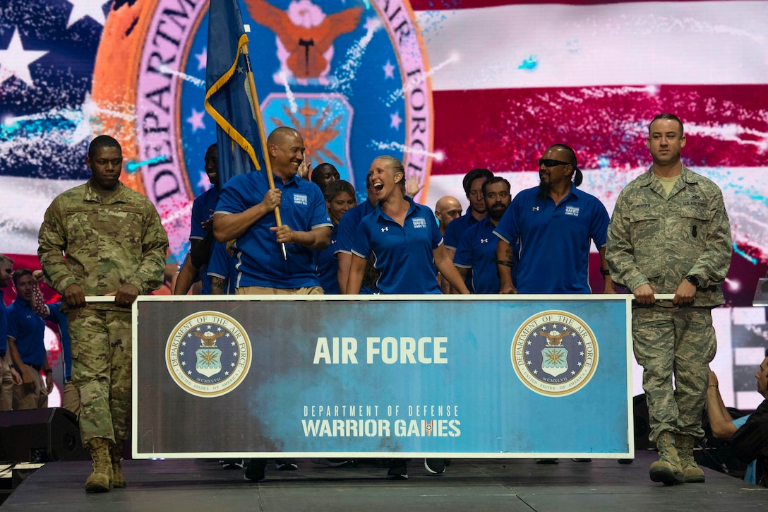 Air Force athletes in team uniforms smile behind a Warrior Games team sign, as one holds up a service flag.