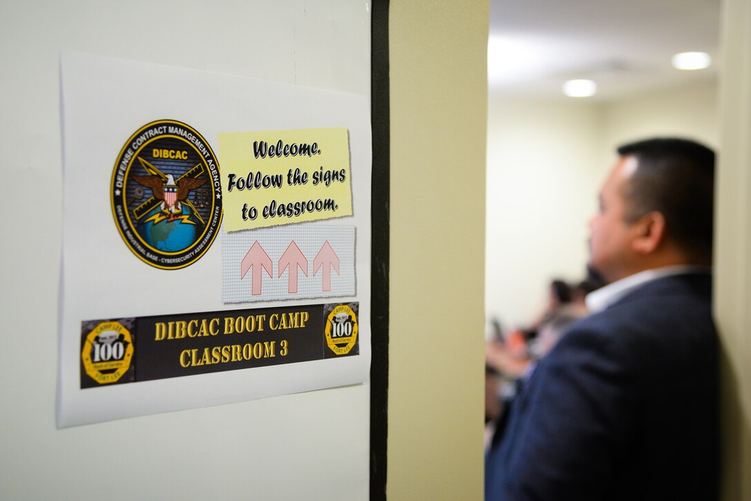 A man stands near a door, the door has a sign that says Welcome, follow the signs to the classroom. DIBCAC Boot Camp.