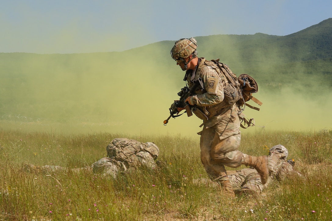 A soldier wearing a combat uniform and carrying a rifle runs behind two other soldiers lying in a field providing cover as smoke spreads.