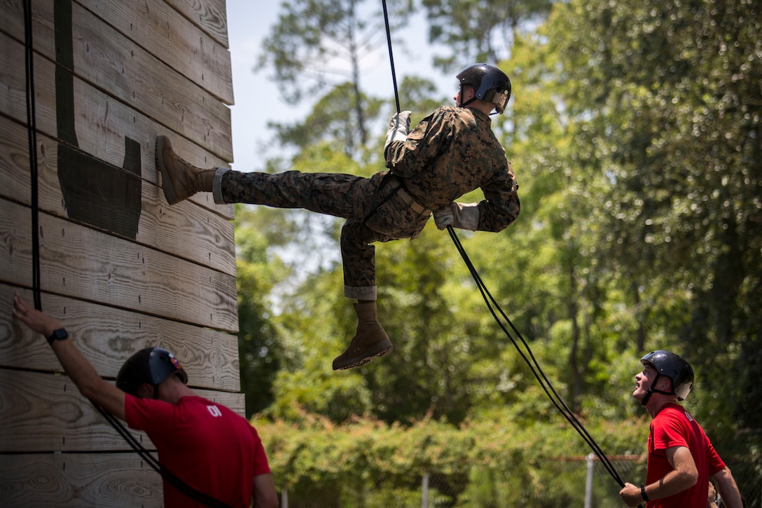 A Marine wearing a harness, a helmet and gloves rappels on a wall as others support and look on.