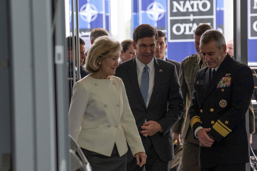 Acting Defense Secretary Mark T. Esper talks with officials while walking through a doorway with NATO signage behind them.