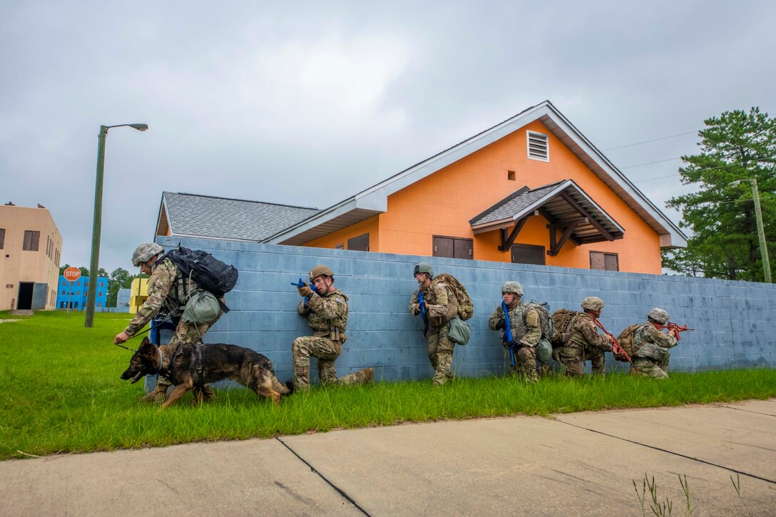 Soldiers stand and kneel in a line against a wall; one soldier holding a dog by a leash.