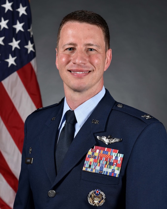 Official photo of white male Air Force colonel dressed in blues posed in front of American Flag.