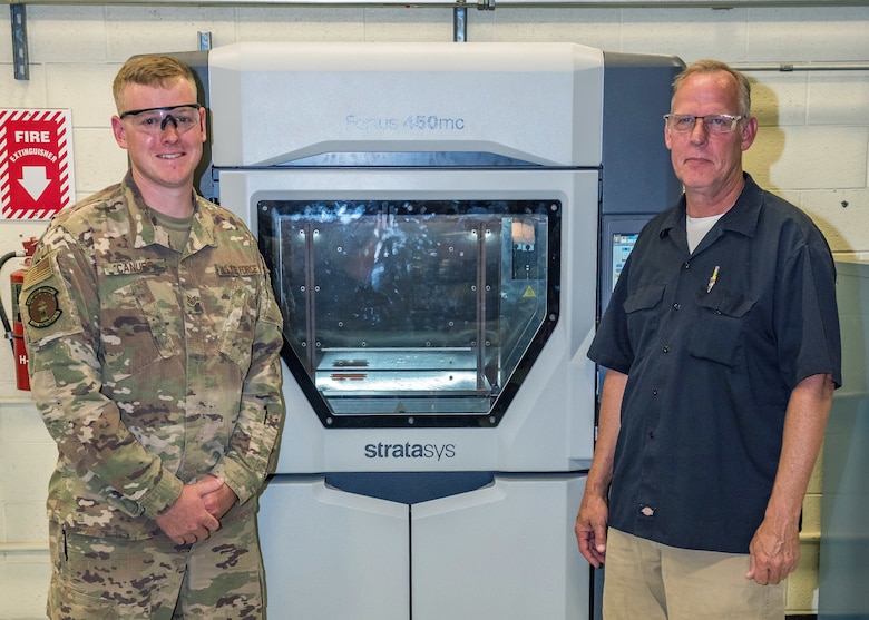 Staff Sgt. Cameron Canupp and Steven Conway, both of 412th Maintenance Squadron, pose for a photo in front of a 3D printer at Edwards Air Force Base, California. The 3D printer can be used to manufacture hard-to-find parts and helps cut down on time maintenance time and costs. (U.S. Air Force photo by Matthew Williams)