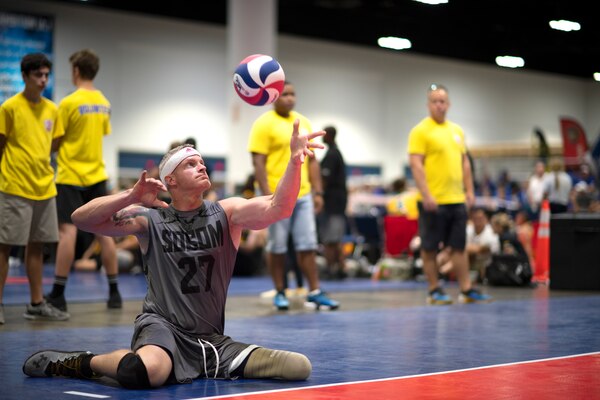 A Wounded Warrior sits on the floor of a volleyball court prepares to serve a ball during a sitting volleyball match at the 2019 DoD Warrior Games in Tampa, Florida