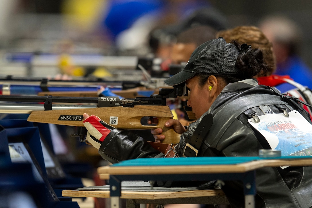 A woman aims a rifle while seated during competition.