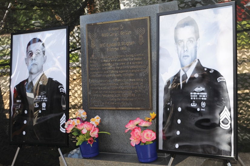 Two large portraits of soldiers in dress uniform are propped up beside a plaque honoring them. Flowers sit at the base of the plaque.