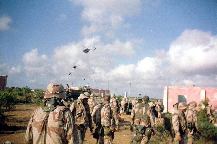 Several soldiers in desert camouflage look into the distant sky, where four Black Hawk helicopters are flying.