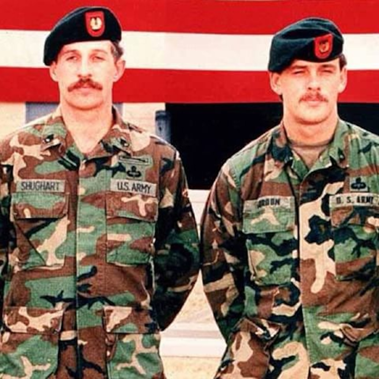 Two Delta Force soldiers wearing camouflage and black hats with red logos stand with their hands behind their backs in front of an American flag.