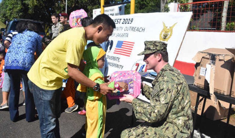 U.S. Navy Petty Officer 1st Class Nickolas Czyzewski, an analyst with the U.S. Indo-Pacific Command Augmentation Team-Indonesia, gives a backpack to a child during the Cope West 19 open house at Sam Ratulangi International Airport, Manado, Indonesia June 22, 2019.  During the event, a team of U.S. Embassy-Jakarta members set up a small kiosk showcasing U.S.-Indonesian 70th anniversary bilateral relations items and educational handouts for children. The team handed out educational books, back packs, writing supply materials, in commemoration of diplomatic relations between the U.S. and Government of Indonesia. (Courtesy photo)