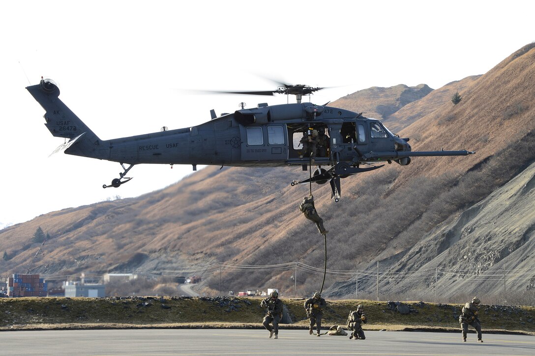 Coast Guardsmen climb down a rope hanging out of a helicopter onto the ground.