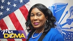 Picture of a smiling lady wearing a blue blouse standing in front of the American flag