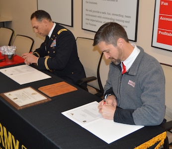 Two men sitting at a table sign documents. The man on the left is wearing dress blue Army uniform and the man on the right is wearing a grey zip up fleece.