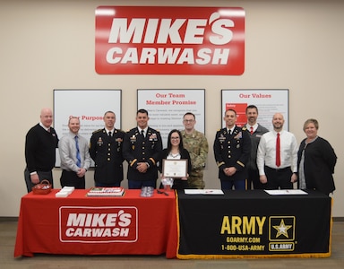 Service members and staff from Mike’s Car Wash gathering around a partnership table with the US Army logo on the right side and a Mikes Car Wash logo on the left side. There are ten people total in the photo. Soldiers are in dress blues and multicam. Other men are wearing shirts and ties and the woman are dressed in work attire. In the background there is a big red Mike’s Car Washing sign with their values and members promise posters underneath it.