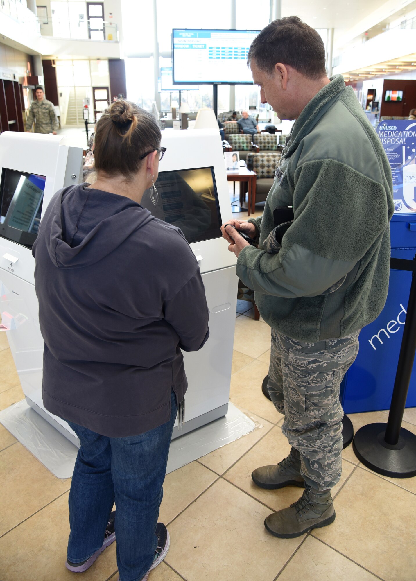 Teresa Larios, pharmacy technician with the 72nd Medical Support Squadron, assists Col. Cliff Altizer, Air Force Sustainment Center Inspector General’s Office, with the new Q-Flow machines inside the lobby of the 72nd Medical Group. The Q-Flow kiosks allow pharmacy customers to check-in to pick up their prescriptions and will show them on the large screens in the waiting area what their wait time is. (U.S. Air Force photo/Kelly White)