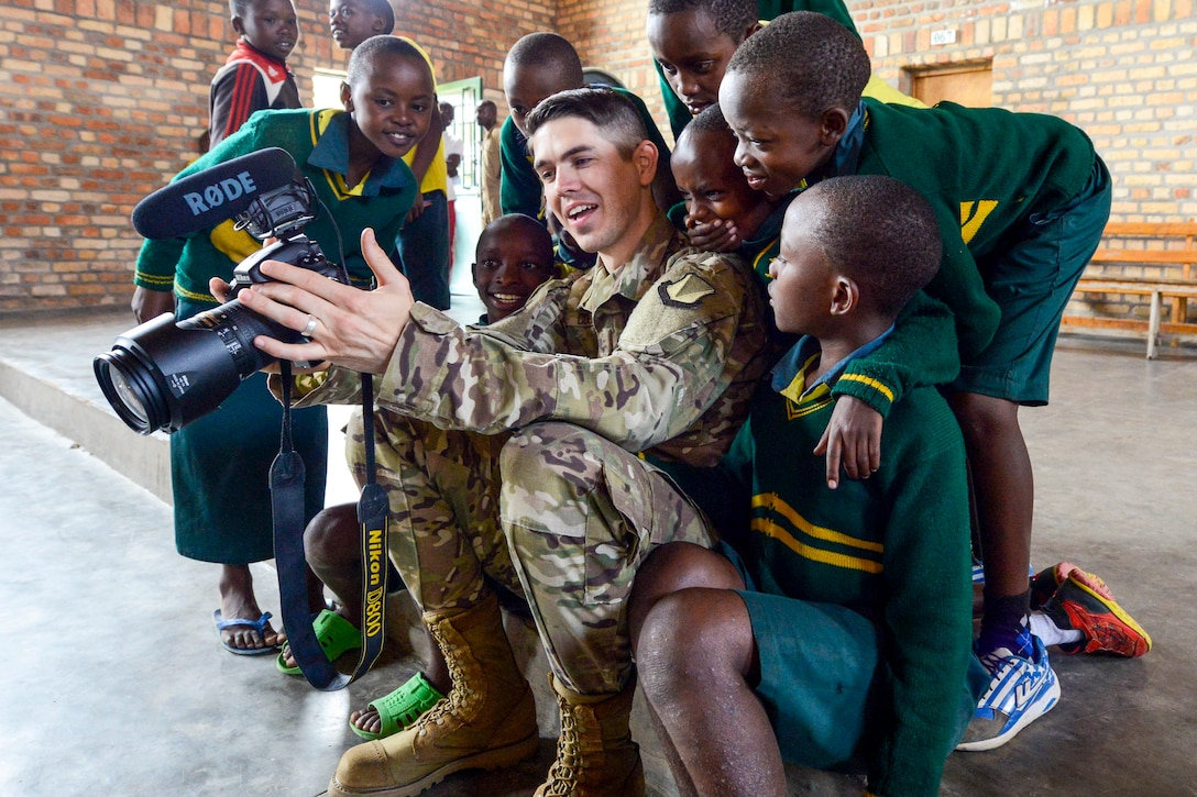 An airman sitting on a floor shows video to a group of smiling boys huddling around him.