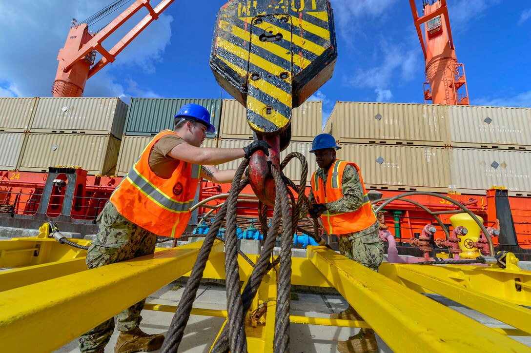 Two sailors attach cables to a crane on a pier.