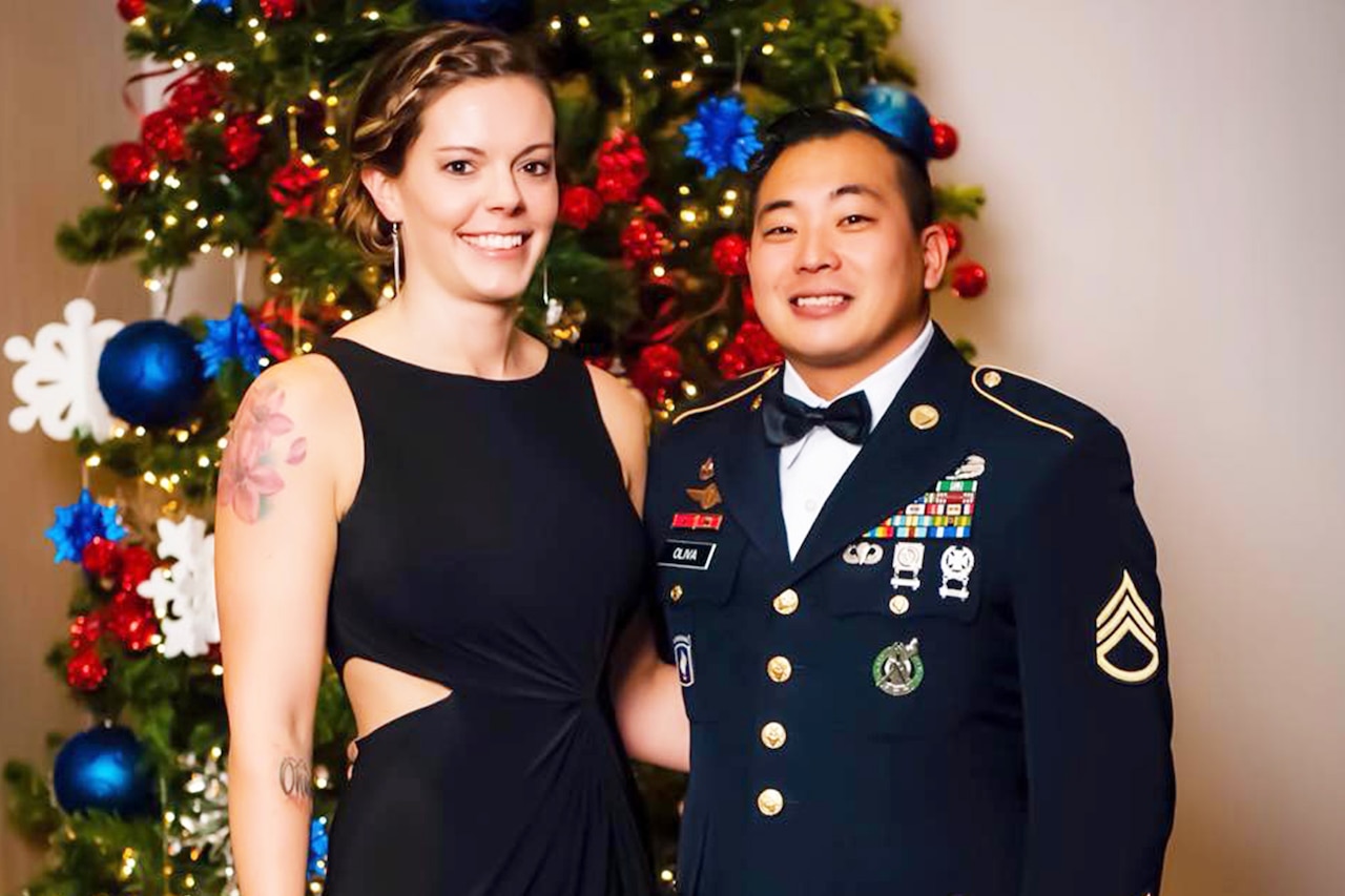 Soldier in uniform stands with wife in front of Christmas tree.