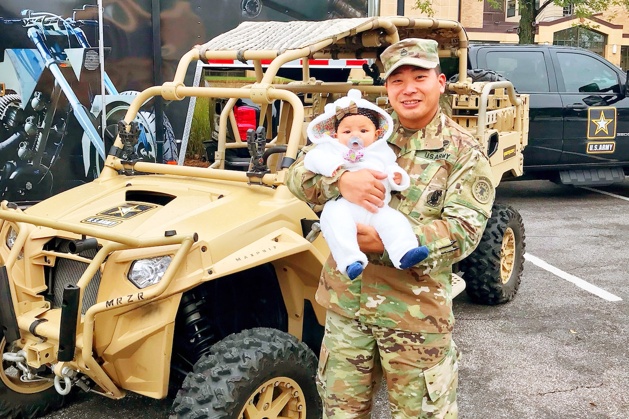 Soldier in uniform holds baby in front of all-terrain vehicle