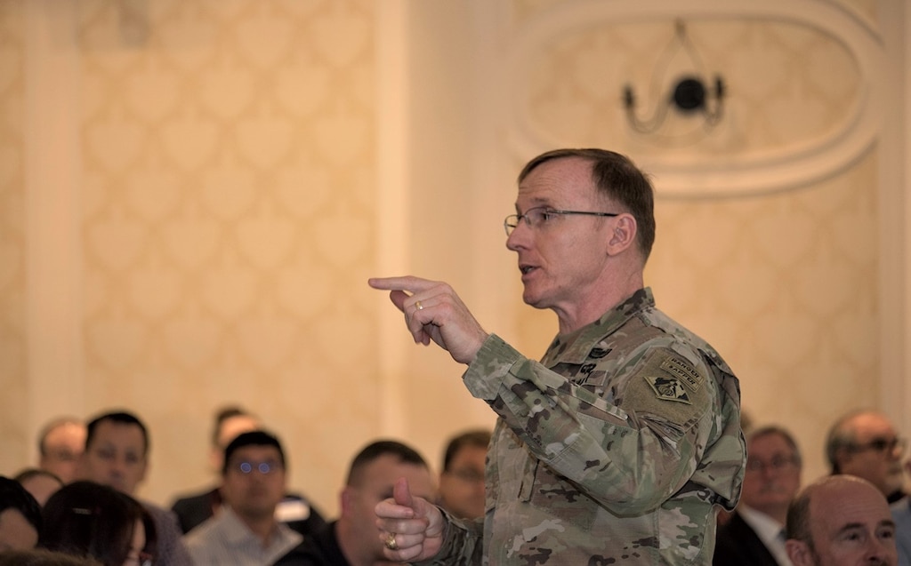 Colonel John Baker stands in front of a group of people, with his hand pointed outward.