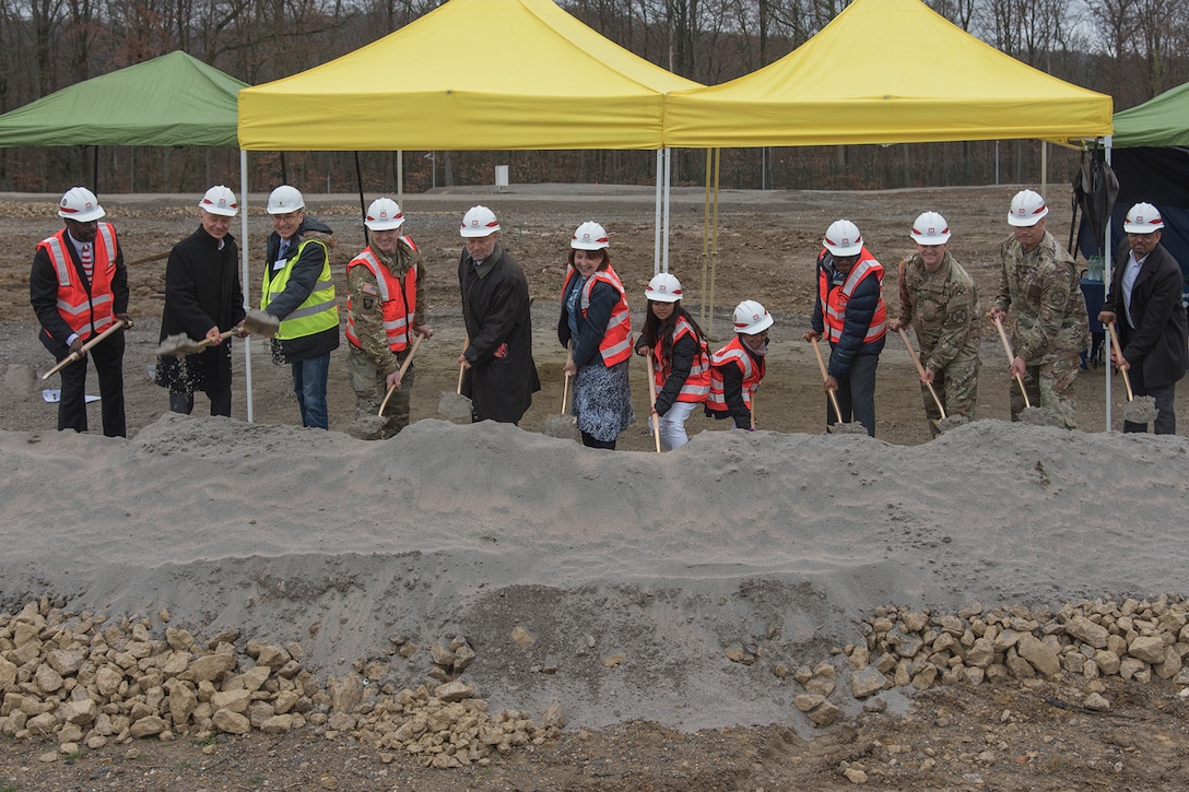 A group of people stand in a line with hard hats and orange safety vests, shoveling dirt.