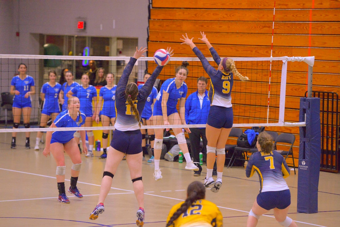 Women volleyball players jump up to block a ball coming from the other side of the net.