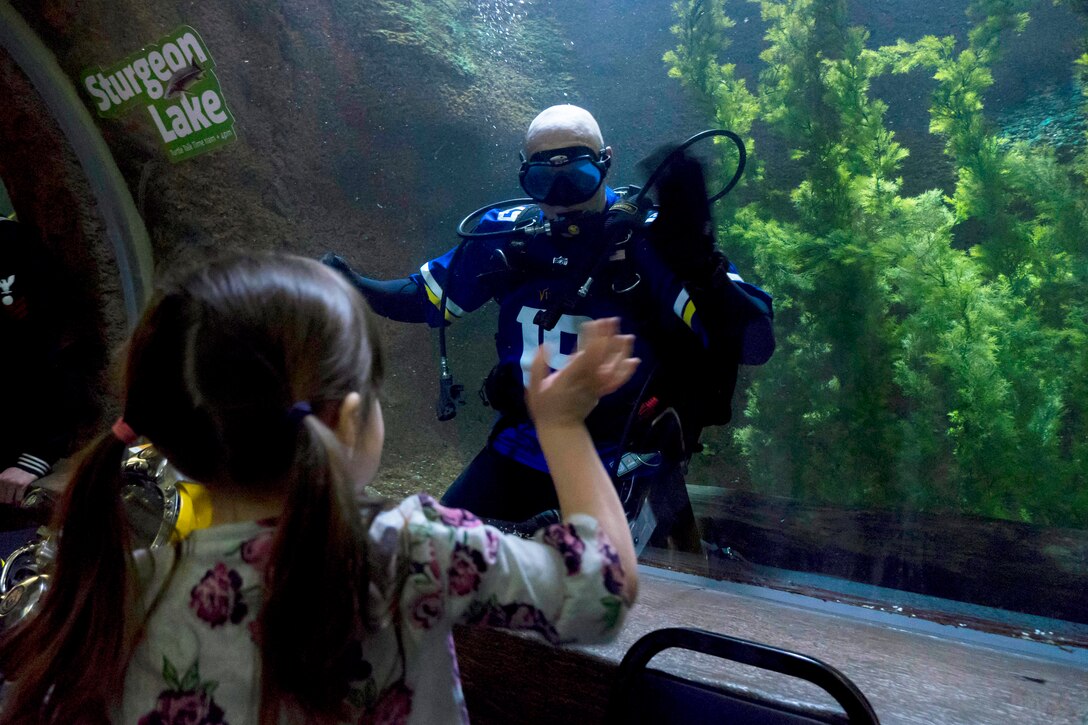 A young girl waves to a diver swimming in an aquarium.