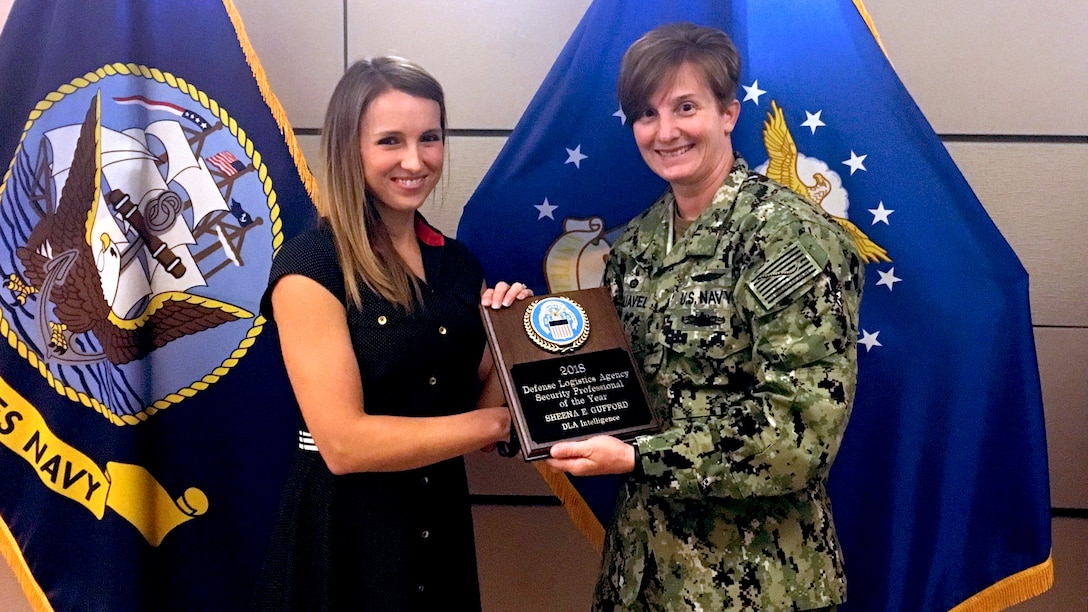 DLA Intelligence Indo-Pacific Security Specialist Sheena Gufford, left, receives an award plaque from DLA Indo-Pacific Commander Navy Capt. Kristin Acquavella, right, in front of flags.