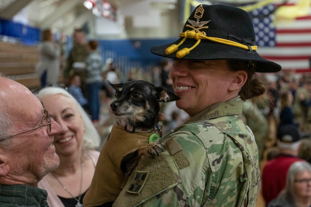 A smiling soldier holds a small dog as the soldier's parents look on, smiling.