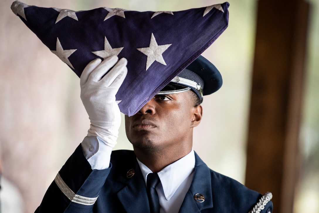 An airman looks at a folded American flag as he holds it in front of his face.