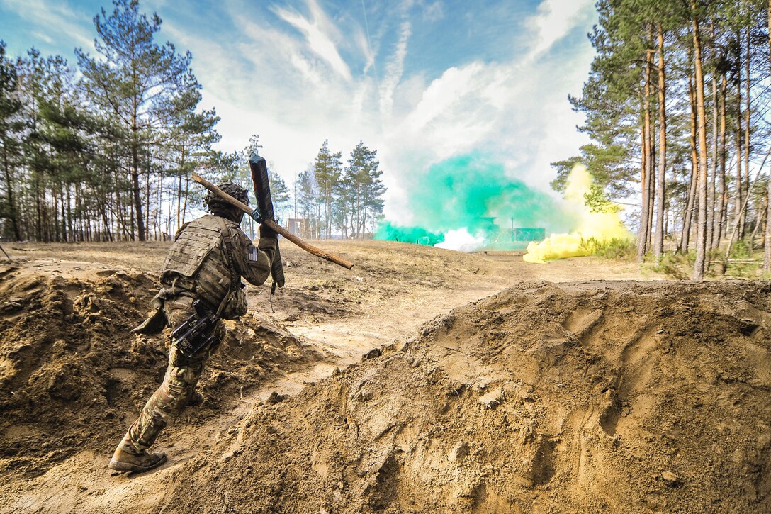 A soldier holding two pieces of wood walks in between two dirt mounds towards green and yellow smoke in the distance