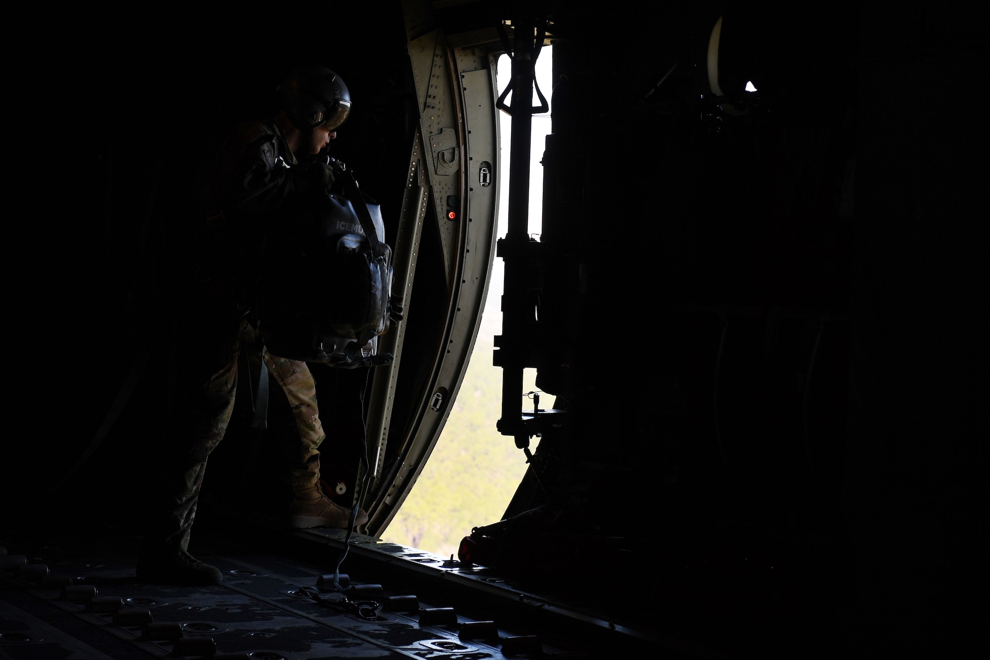 A man holds a backpack in front of an open door of a C-130J as it flies through the air.