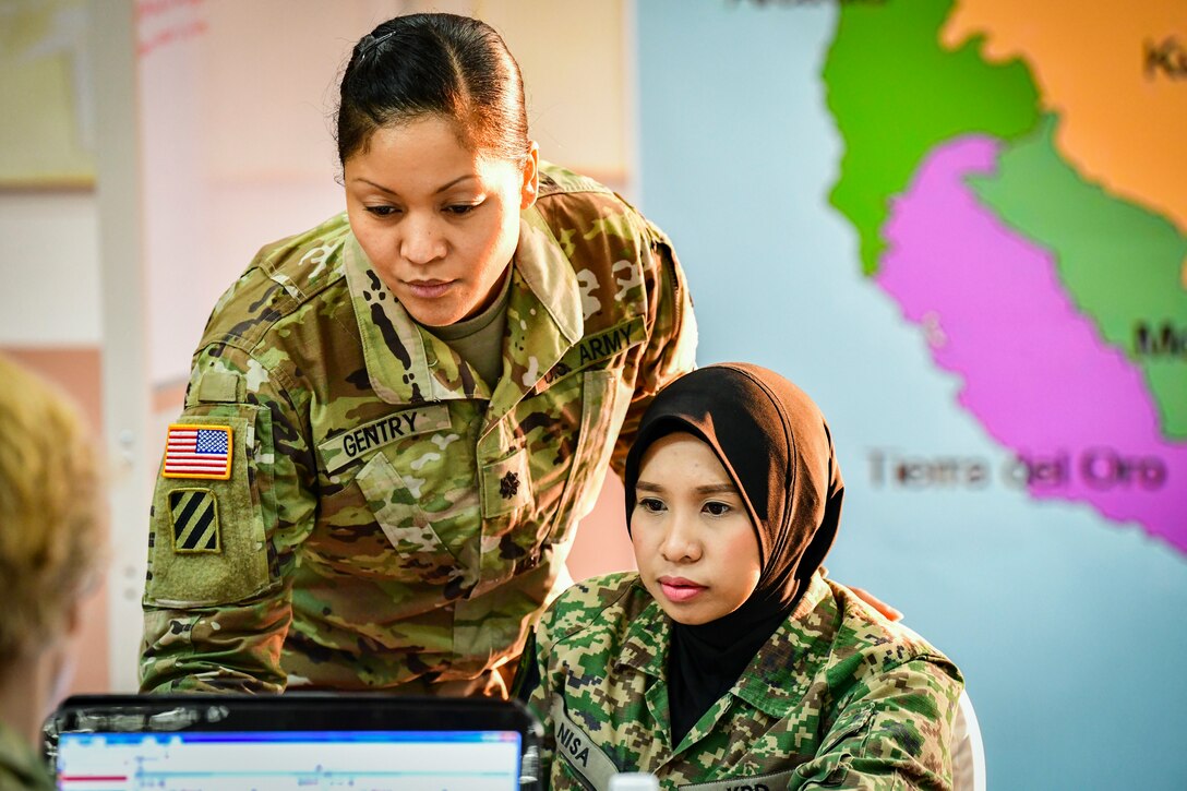 A soldier stands over the shoulder of a soldier sitting behind a computer