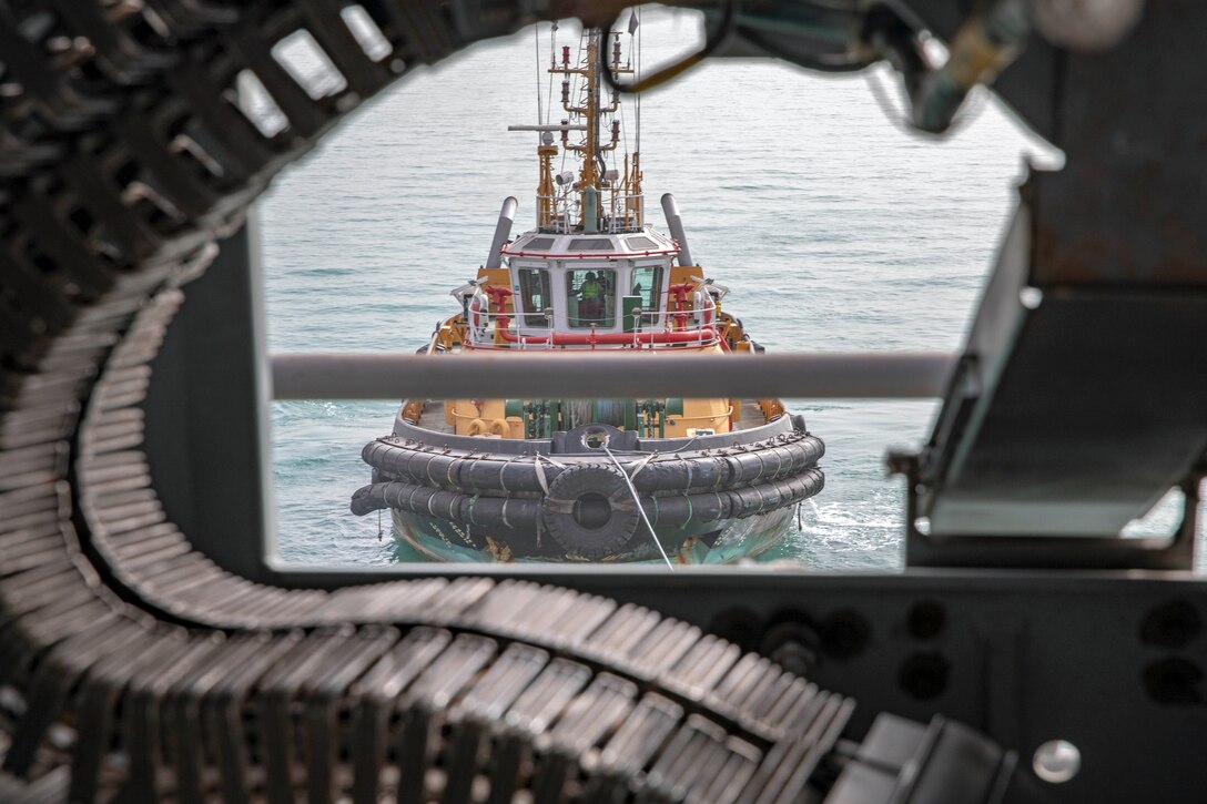 The curve of a machine gun ammunition belt on a ship frames a view of a nearby tugboat in water.