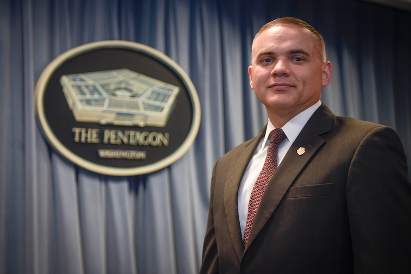 A man in a suit stands to the right of a Pentagon sign.