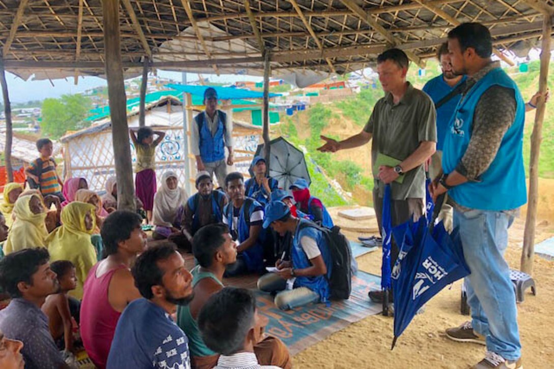 Two standing men talk to several men seated under a hut.