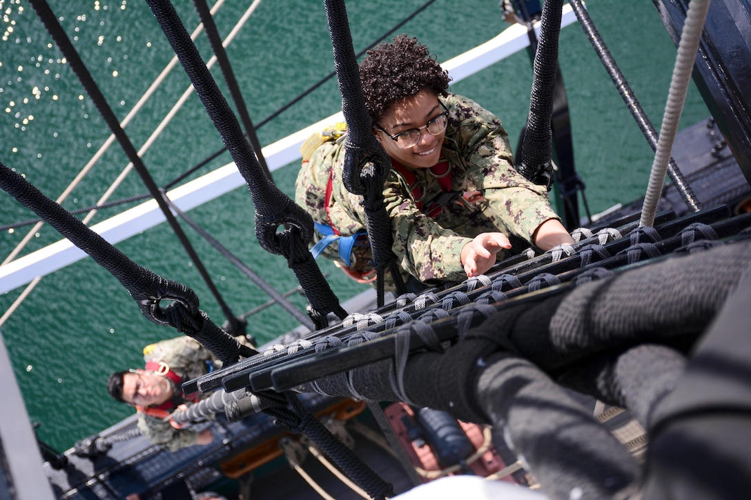 A sailor climbs the mizzenmast of a ship while another sailor watches from below.