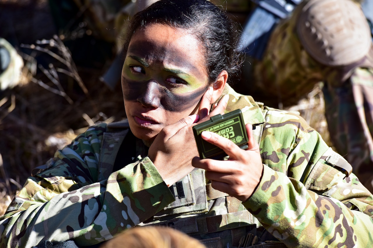 A soldier paints her face green and black while holding a small mirror.
