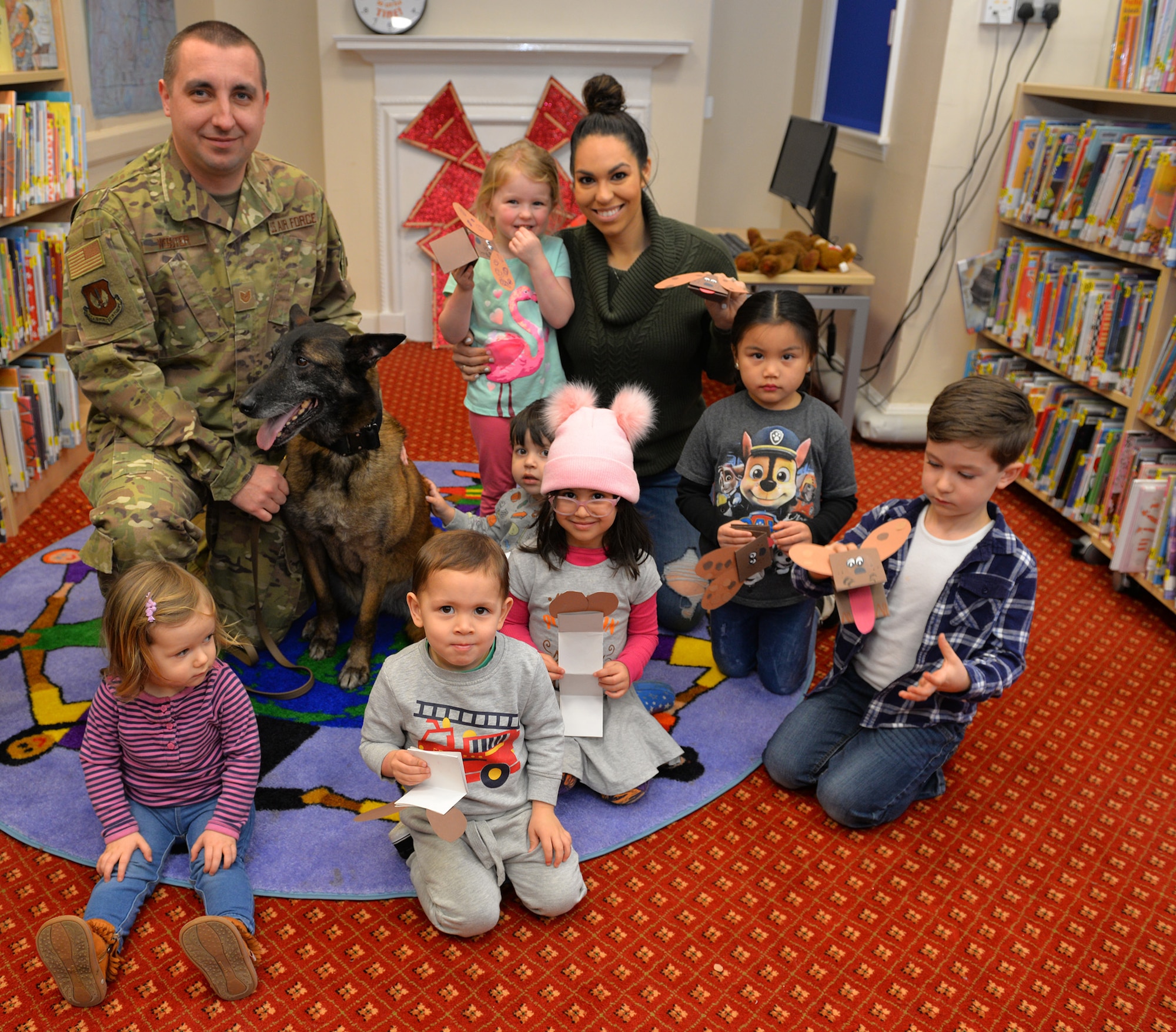 U.S. Air Force Tech. Sgt. Kelly Webster, 100th Security Forces Squadron Military Working Dog kennel master, retired MWD Vvonya, and Karina Santos, 100th Force Support Squadron Library programs manager, pose for a photo with Team Mildenhall children after a craft session at the base library on RAF Mildenhall, England, March 13, 2019. To celebrate K9 Veterans’ Day, Webster read a story to children before they made crafts, did coloring and had the chance to pet Vvonya. Webster and his family adopted Vvonya after her retirement in January. (U.S. Air Force photo by Karen Abeyasekere)