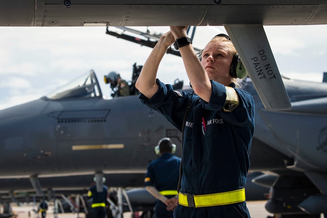 An airman uses a tool to work on a jet with another jet in the background.