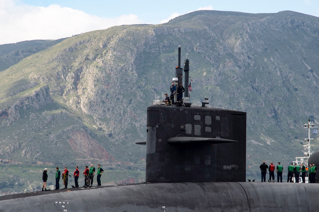 Two groups of people stand on top of a submarine with large hills in the background.