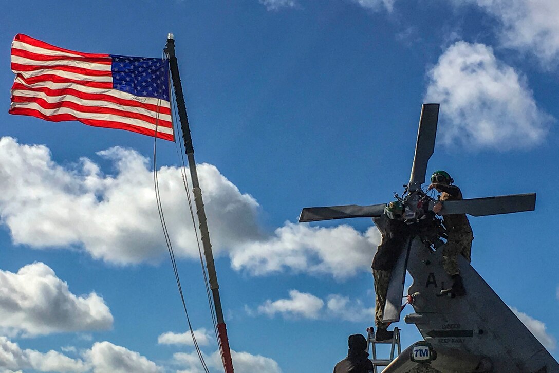 A sailor stands on top of a helicopter while working on it's wing.