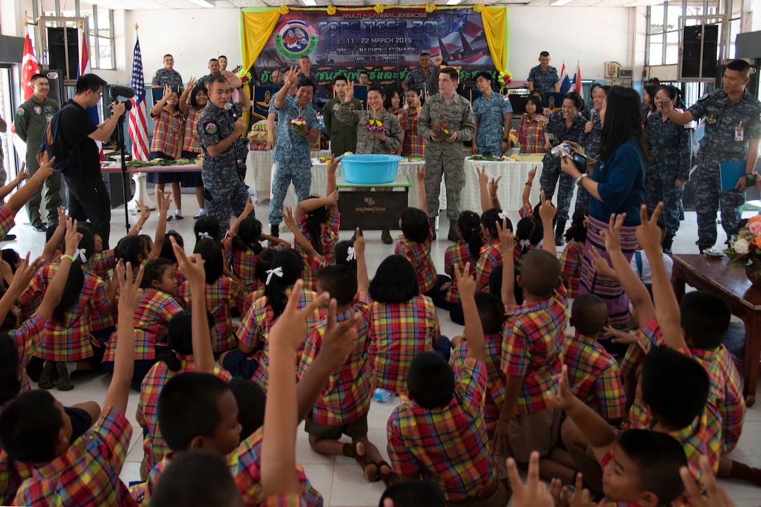 A large group of children raise their hands during a class with some military men standing in front of them.