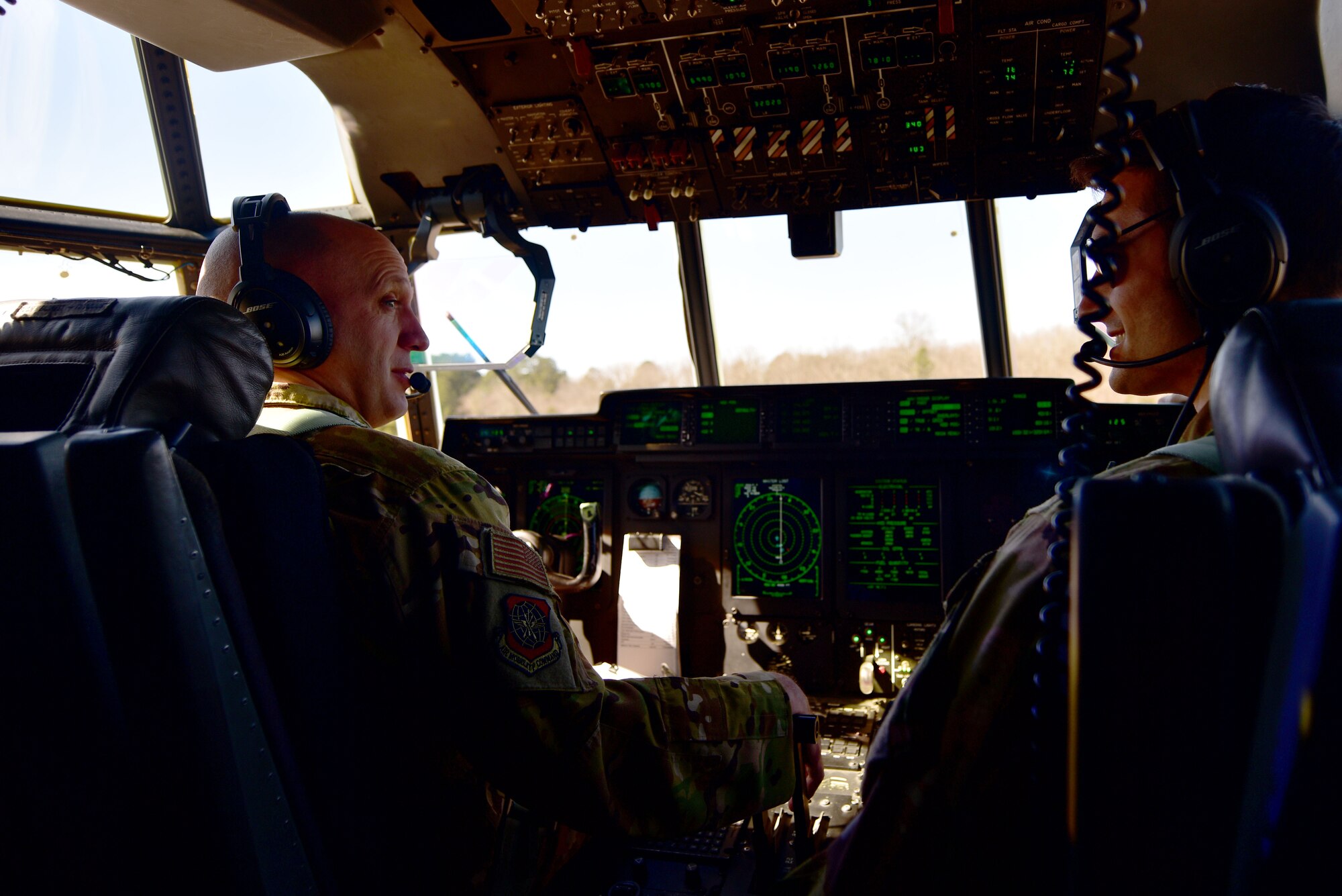 A man wearing a headset in the flight deck of a C-130J talks to another man.