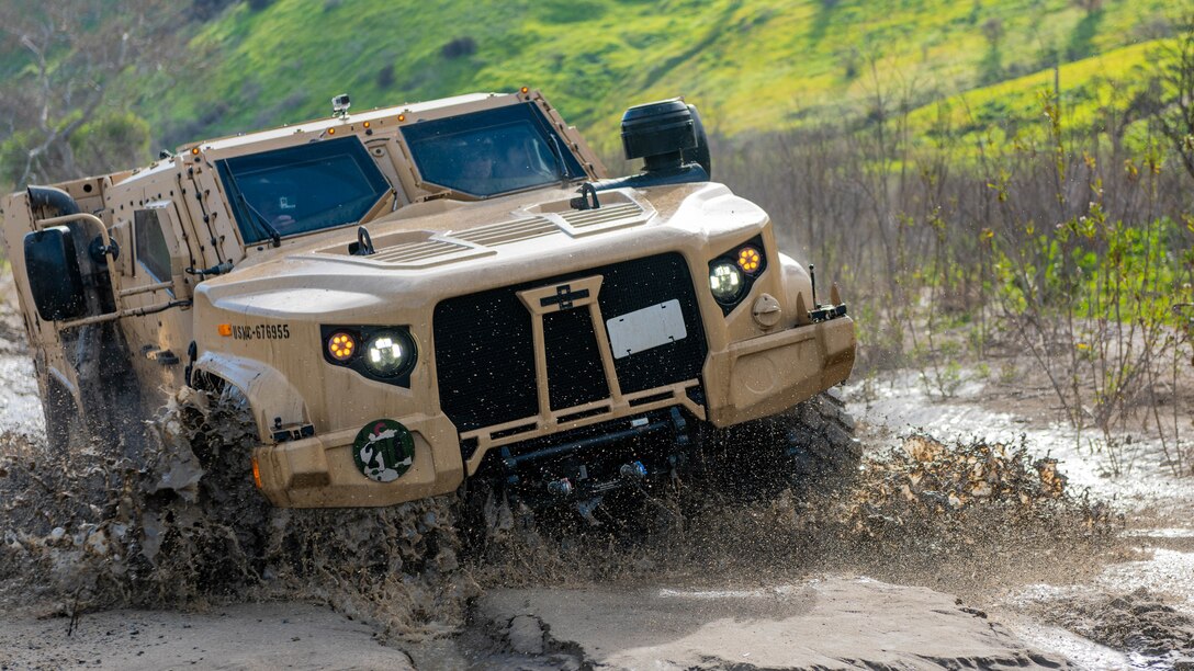 A tactical vehicle splashes through water and mud.